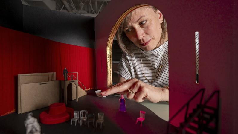 Sara Brown’s hand reaches through a tiny door into a miniature model of a red room with paper people and chairs.
