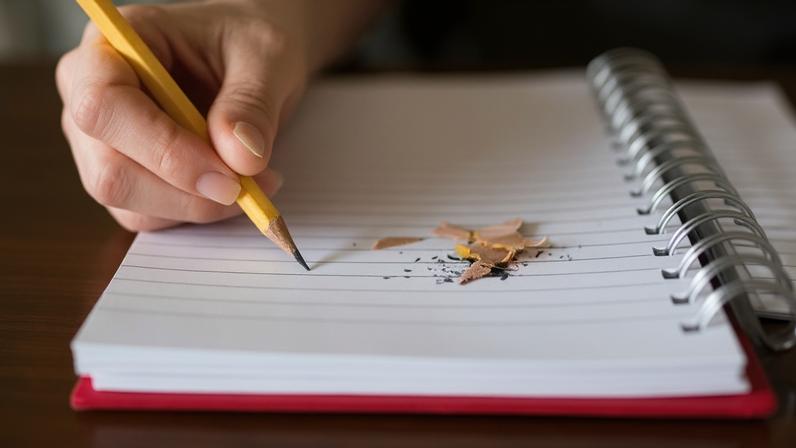 Pessoa escrevendo em um caderno. Há ponta de lápis em cima da folha