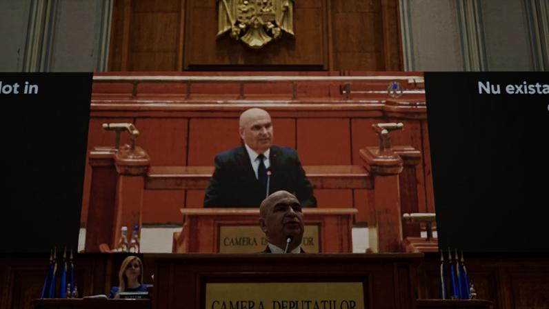 Romanian Prime Minister Ilie Bolojan stands in front of a podium to address parliament. A large screen airing his speech live displays behind him.