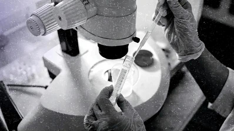 Imagem focada nas mãos de um cientista manipulando uma proveta diante de um microscópio.