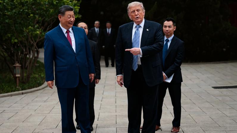 Trump and Xi walk side by side along a footpath, wearing dark business suits and ties, with aides following along in the background. Trump is pointing one finger at Xi, mouth open as he speaks.