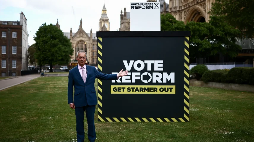 Reform UK leader Nigel Farage poses in front of a sign reading, “Vote Reform, Get Starmer Out.” He is wearing a suit, and his left arm is outstretched.