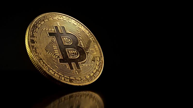 Bitcoin coin with dramatic purple lighting on dark reflective surface suggesting price decline
