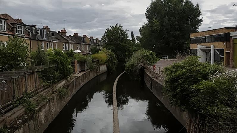 The river Wandle runs past houses in Wandsworth on August 24, 2020 in London, England. The river is in the middle and is enclosed by concrete banks on either side. On the left is a row of terraced houses and on the right is a modern brick and glass 1960s style building. There is some vegetation on the river banks.