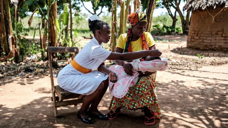 A midwife in a white uniform with an orange belt sits outdoors, leaning forward to examine a newborn baby held by a woman in a yellow shirt and colorful patterned skirt. They are seated in a sunlit, dirt clearing surrounded by trees and a traditional thatched-roof building in the background.
