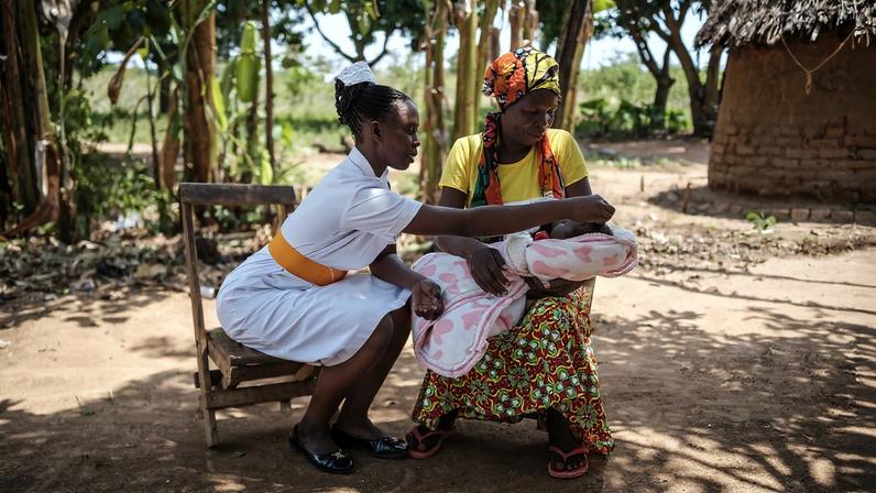 A midwife in a white uniform with an orange belt sits outdoors, leaning forward to examine a newborn baby held by a woman in a yellow shirt and colorful patterned skirt. They are seated in a sunlit, dirt clearing surrounded by trees and a traditional thatched-roof building in the background.