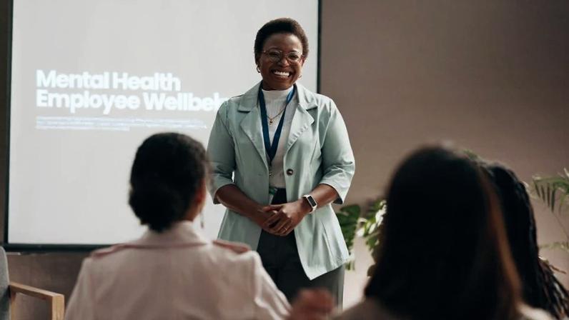 A NR-1 entra em vigor e torna saúde mental obrigação nas empresas. Estudo da Yale aponta alta de 25% na produtividade com apoio psicológico. Happy woman, presentation and meeting with group for mental health program or employee wellbeing at office. Female person, speaker or crowd with smile for seminar, conference or social workshop