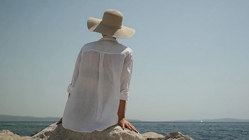 A woman sat in the sun wearing a loose shirt and sun hat