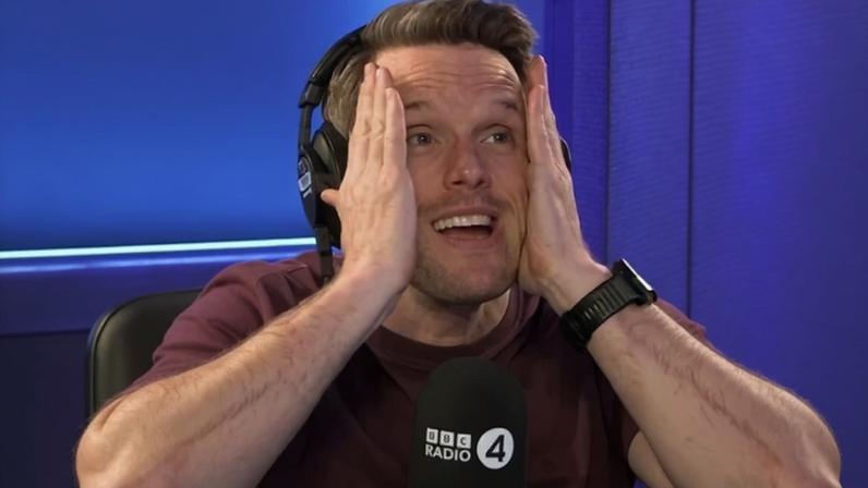 Greg Foot clasps his head in his hands and looks worried. He is wearing a brown T-shirt and is sat behind a Radio 4 mic.