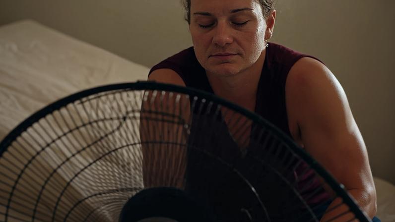 A woman sits on a bed in front of a fan