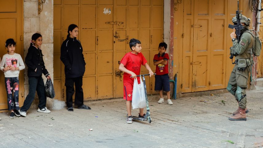 An armed Israeli soldier stands guard near children standing by closed shop doors.