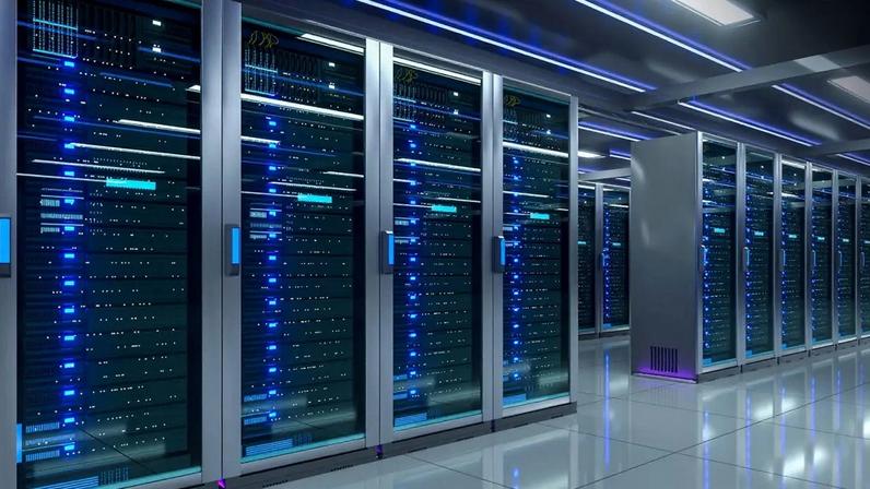 Corredor de um data center moderno com fileiras de racks de servidores, exibindo luzes azuis piscando, refletidas no piso brilhante. O ambiente é escuro, com iluminação de teto embutida e linhas de luz azul no alto. No canto superior direito, há um círculo branco com a letra "S" em preto