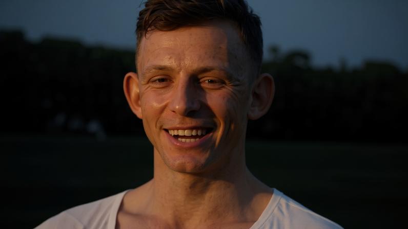 Homem jovem, branco, com cabelo curto e escuro, sorrindo amplamente com os dentes à mostra, vestindo uma camiseta clara, em um fundo desfocado de árvores e céu