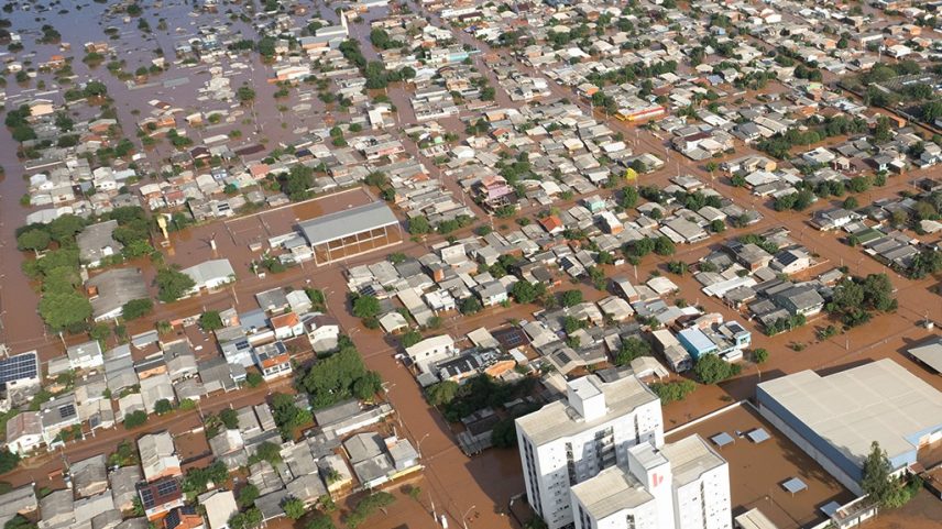 Vista aérea de uma cidade inundada, com casas e ruas submersas em água barrenta. Telhados cinzas e marrons se destacam na paisagem alagada, com algumas árvores e edifícios maiores, como um ginásio e prédios brancos, parcialmente visíveis. A extensão da inundação é vasta, cobrindo grande parte da área urbana.