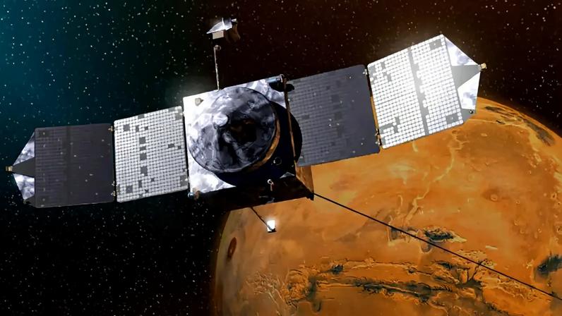 Sonda espacial cinza com painéis solares retangulares estendidos, orbitando o planeta Marte, que aparece em tons de laranja e marrom com crateras visíveis, em um fundo de espaço escuro com estrelas