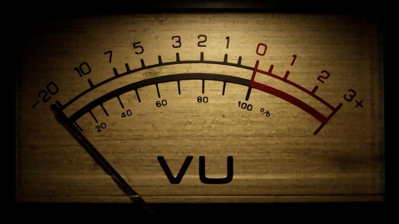Medidor de VU analógico com ponteiro vermelho marcando a zona de sobrecarga, indicando volume alto. A escala superior vai de -20 a +3, e a inferior de 20% a 100%