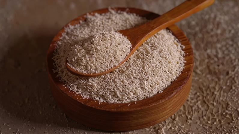Fibras de psyllium em uma tigela de madeira com uma colher de pau cheia, e folhas verdes de manjericão ao fundo