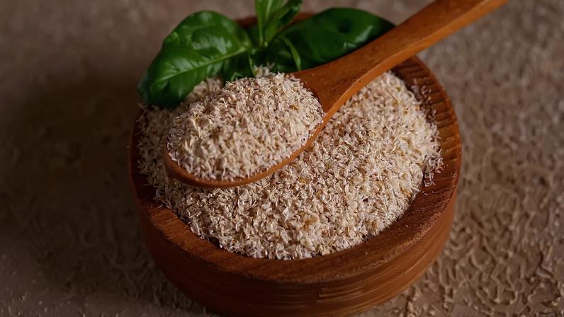 Fibras de psyllium em uma tigela de madeira com uma colher de pau cheia, e folhas verdes de manjericão ao fundo