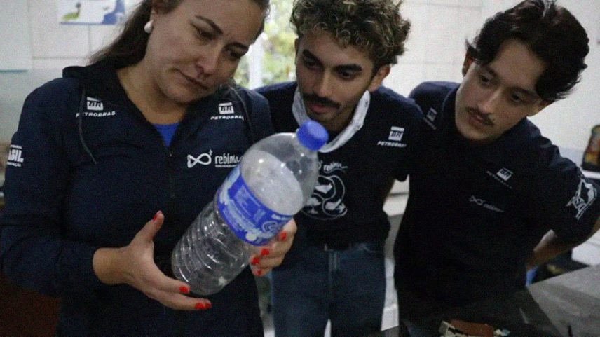 Mulher de blusa azul escura com logo Petrobras e Rebinma, unhas vermelhas, segura uma garrafa plástica transparente com líquido branco. Dois homens jovens, um com cabelo cacheado e outro com bigode, ambos de camisetas escuras com logos Petrobras e Rebinma, observam a garrafa atentamente