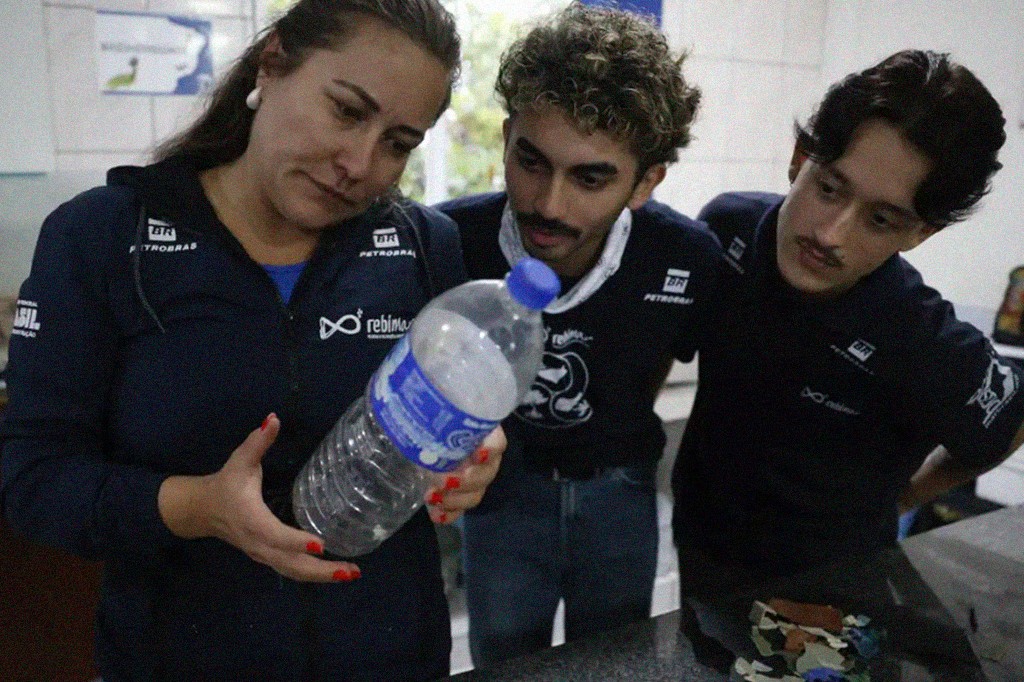 Mulher de blusa azul escura com logo Petrobras e Rebinma, unhas vermelhas, segura uma garrafa plástica transparente com líquido branco. Dois homens jovens, um com cabelo cacheado e outro com bigode, ambos de camisetas escuras com logos Petrobras e Rebinma, observam a garrafa atentamente