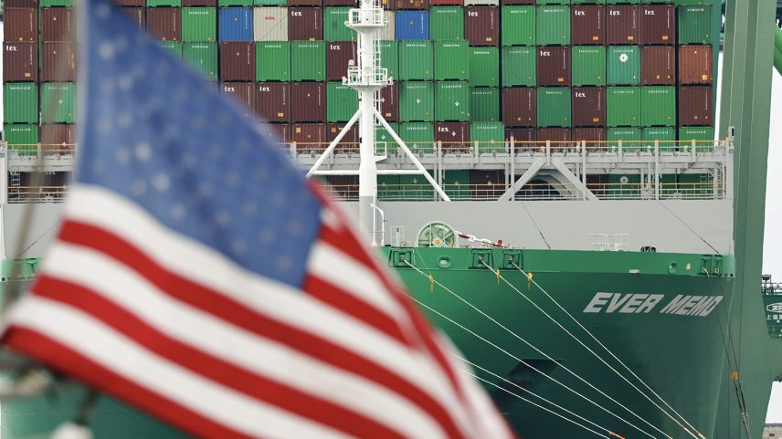A close-up shot focuses on a massive cargo ship loaded with shipping containers, framed by a blurred American flag in the foreground. The flag, with its red and white stripes and blue field of white stars, is positioned on the left side of the frame and is heavily out of focus. Behind it, the green bow of a large container ship is sharply detailed, bearing the partial white text "EVER MEMO." Tied lines stretch from the ship's bow toward the right. Towering above the hull are rows of stacked shipping containers in various colors, predominantly green and brown, with some displaying white text like "tex." A white ship mast stands near the center of the vessel against the cargo backdrop.