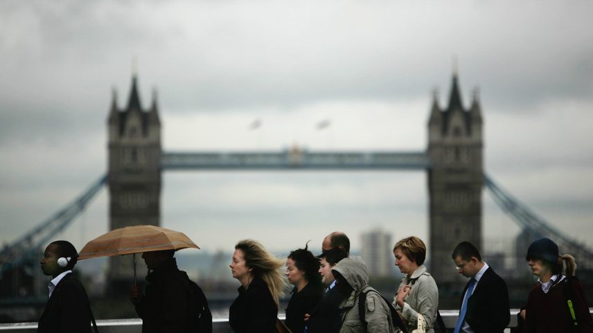 A line of commuters walking in profile against a cloudy sky, with a large, historic suspension bridge featuring two prominent towers softly blurred in the background. One person holds a brown umbrella.