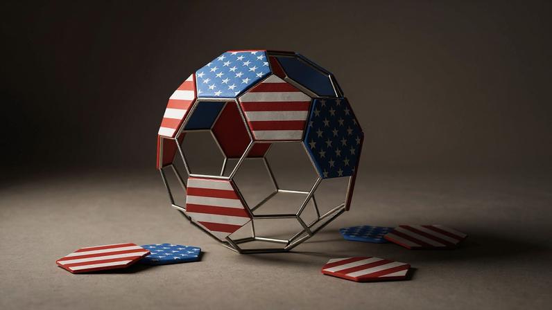 An illustration of a soccer ball wireframe structure resting on a plain, light beige surface. Several of the hexagonal and pentagonal panels are covered with the stars-and-stripes design of the American flag, while other panels have fallen off and are scattered on the ground around the base of the ball, leaving the metal frame partially exposed.