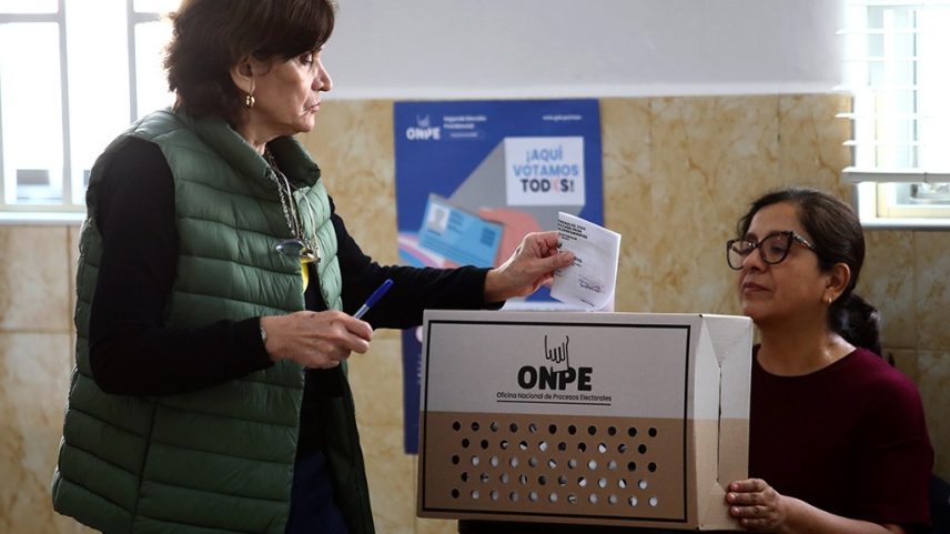 Mulher de colete verde escuro e blusa preta depositando um voto em uma urna eleitoral de papelão, enquanto outra mulher de óculos e blusa vinho observa. A urna tem o logo da ONPE e um cartaz azul ao fundo diz ¡Aquí votamos todos!.