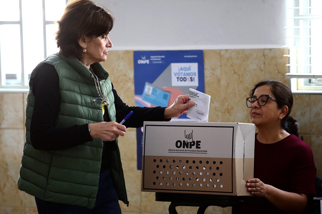 Mulher de colete verde escuro e blusa preta depositando um voto em uma urna eleitoral de papelão, enquanto outra mulher de óculos e blusa vinho observa. A urna tem o logo da ONPE e um cartaz azul ao fundo diz ¡Aquí votamos todos!.