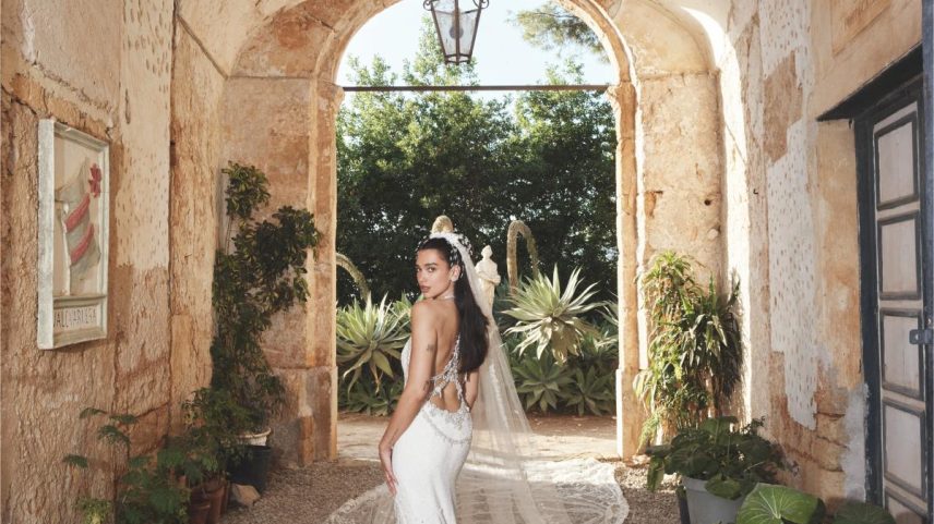 Dua Lipa in a white wedding gown with a flowing veil, standing in an arched stone pathway.