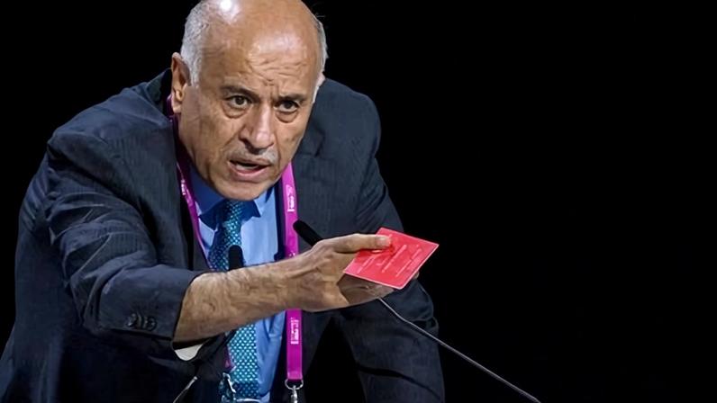 Jibril Rajoub leans forward at a podium holding a red card, speaking at a FIFA event
