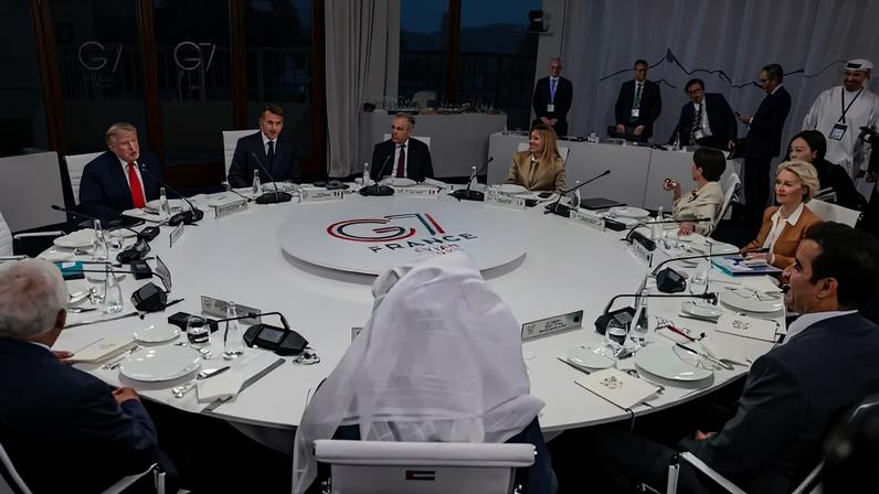 Líderes mundiais sentados em uma mesa redonda branca com o logo G7 France Evian 2021 no centro. Donald Trump e Emmanuel Macron estão à esquerda, enquanto outros delegados, incluindo mulheres e um homem com vestes árabes, participam da reunião. Alguns assessores estão de pé ao fundo.