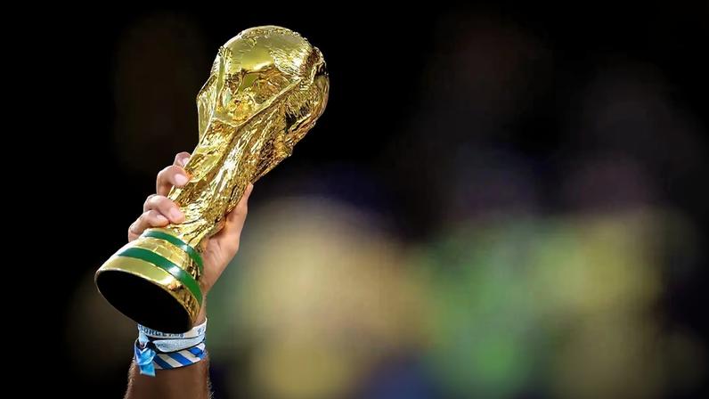 Uma mão segura a taça da Copa do Mundo, dourada com detalhes verdes na base, contra um fundo escuro e desfocado.