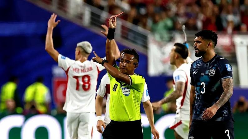 Árbitro de futebol com camisa verde-limão e calção preto, erguendo um cartão vermelho. Ao lado, um jogador de camisa azul escura com o número 3 e outro de camisa branca com o número 18, ambos observando a decisão. O cenário é um estádio de futebol com torcedores ao fundo.