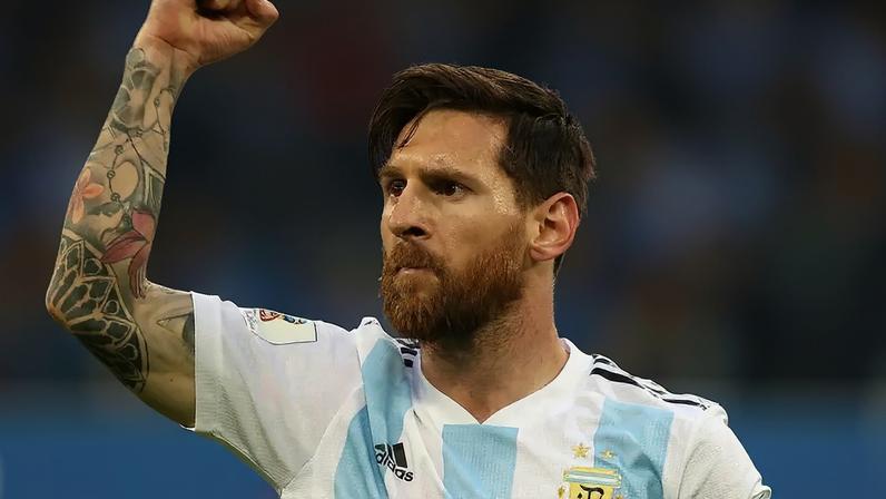 Lionel Messi, com barba ruiva e cabelo castanho, veste a camisa branca e azul da Argentina, erguendo o braço esquerdo tatuado em celebração