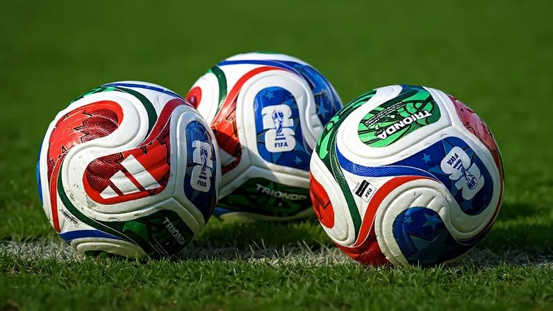 Três bolas de futebol Adidas Brazuca coloridas, com detalhes em vermelho, azul, verde e branco, sobre a grama verde de um campo, próximas a uma linha branca