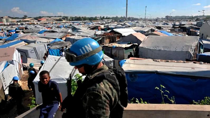 Um soldado da ONU com capacete azul e óculos de proteção, vestindo uniforme camuflado com a bandeira do Brasil no braço, observa um acampamento de refugiados. Tendas de lona cinza e azul se estendem sob um céu claro, com um menino de camisa azul olhando para a câmera em primeiro plano. Ao fundo, mais tendas e algumas construções precárias.