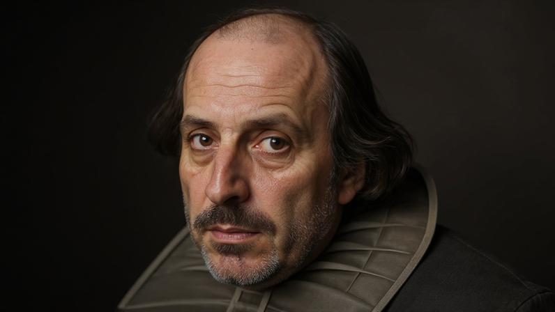 Retrato gravado de William Shakespeare, com cabeça calva, cabelos longos e ondulados nas laterais, bigode e barba rala. Ele usa um colarinho branco plissado e olha diretamente para frente. O fundo é texturizado com linhas cruzadas