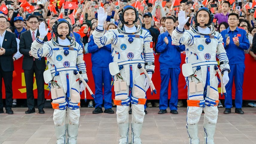 Três astronautas chineses, dois homens e uma mulher, em trajes espaciais brancos e azuis, acenam para uma multidão animada que segura bandeiras da China e de Hong Kong