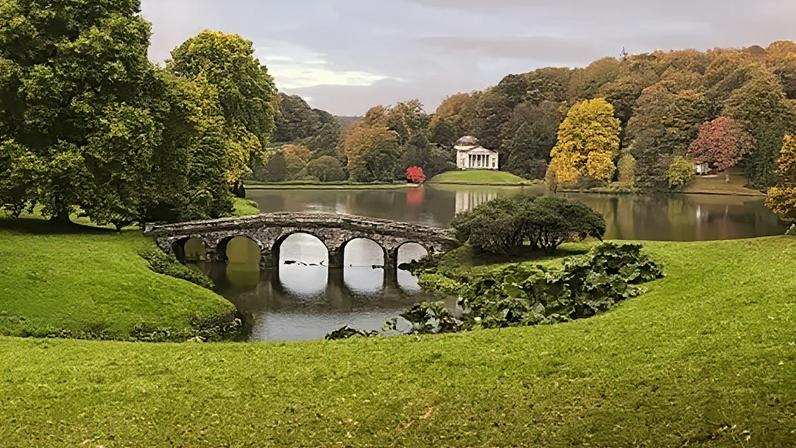 Part of Stourhead's landscape gardens on a cloudy day. Green lawn in front of a lake surrounded by trees. An elegant bridge with arches goes over a narrower part of the lake at the front and in the background is a building designed to look like a classical temple.
