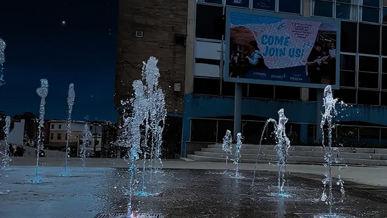 Somerset Council Water jets shoot upwards between paving slabs in a wide open space. The ground is wet due to the jets and a large, run-down building is seen behind.