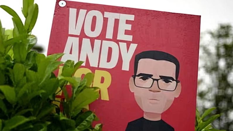 A placard in support of Andy Burnham is partly obscured by plants in the garden of a residential property in Ashton-in-Makerfield