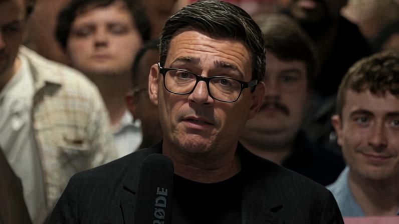 Andy Burnham in glasses and a black t-shirt gestures with his hands while speaking to an audience
