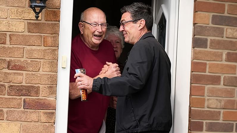 Andy Burnham smiling and clasping a man's hand on a doorstep.