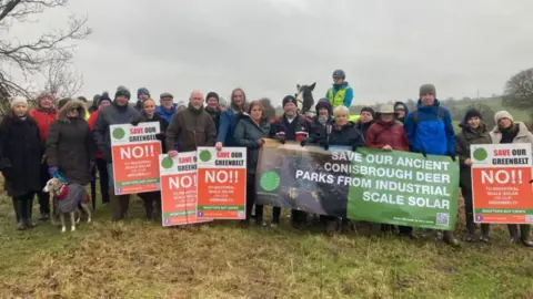 BBC A group of people are stood in a field with white, orange and green placards opposing plans for a solar farm.