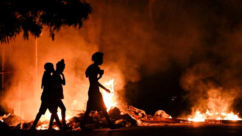The silhouettes of three people are shown at night as they walk past fires.