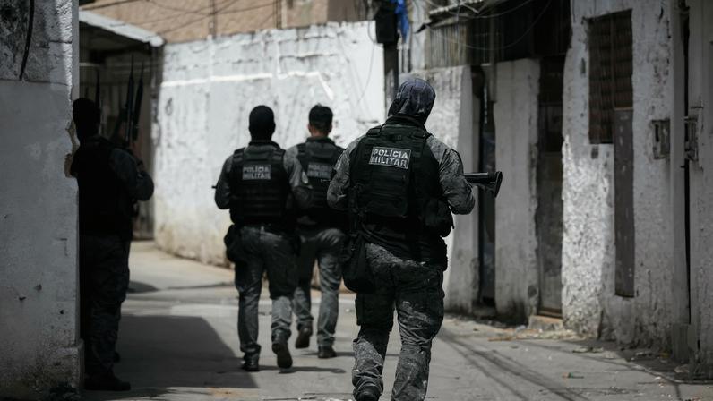About half a dozen people in gray uniform fatigues and bulletproof vests are seen from behind in a narrow street or alley bordered with slightly run-down buildings. Most of the officers hug a wall on the left, but one walks up the middle of the street. All appear to be carrying rifles.