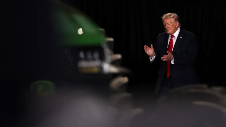 Trump, wearing a black suit and red tie, claps his hands. He is standing next to an out-of-focus green tractor against a dark background.
