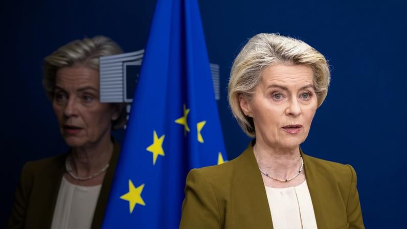 Ursula von der Leyen stands in front of the blue European Union flag and a blue background. Her reflection is seen to the left.