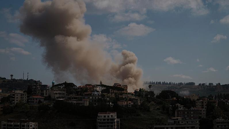 Clouds of smoke billow up from a skyline made up of low- and midrise buildings beneath a hazy blue sky.